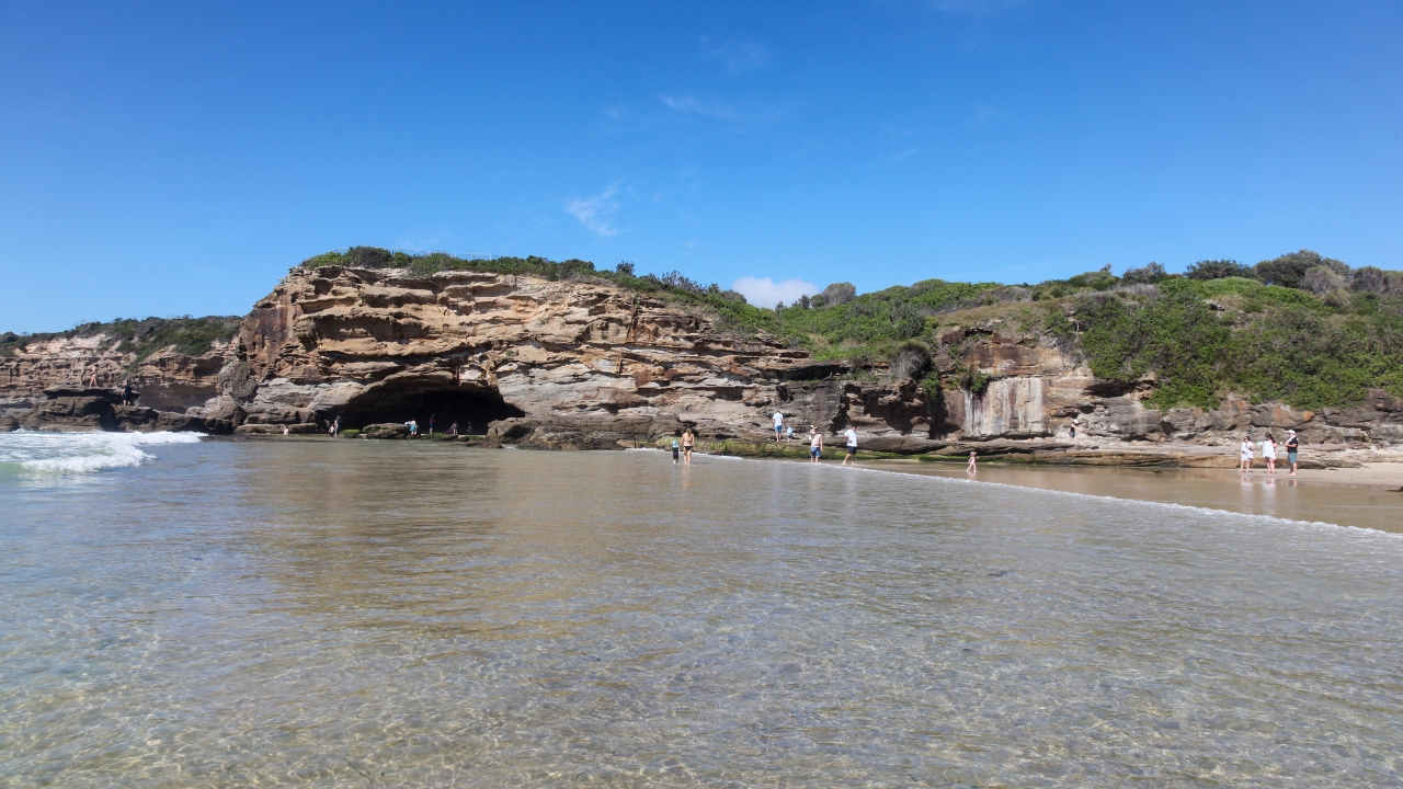 People exploring shallow coastal waters at Caves Beach in Swansea under clear blue sky