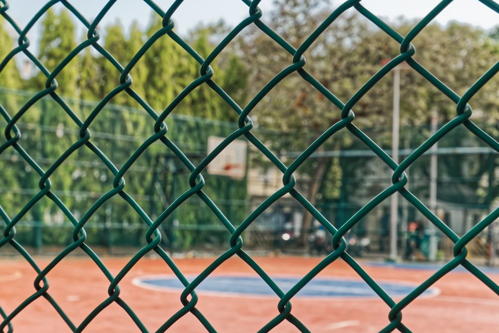 Green chainwire mesh fencing in front of an outdoor basketball court with red surface
