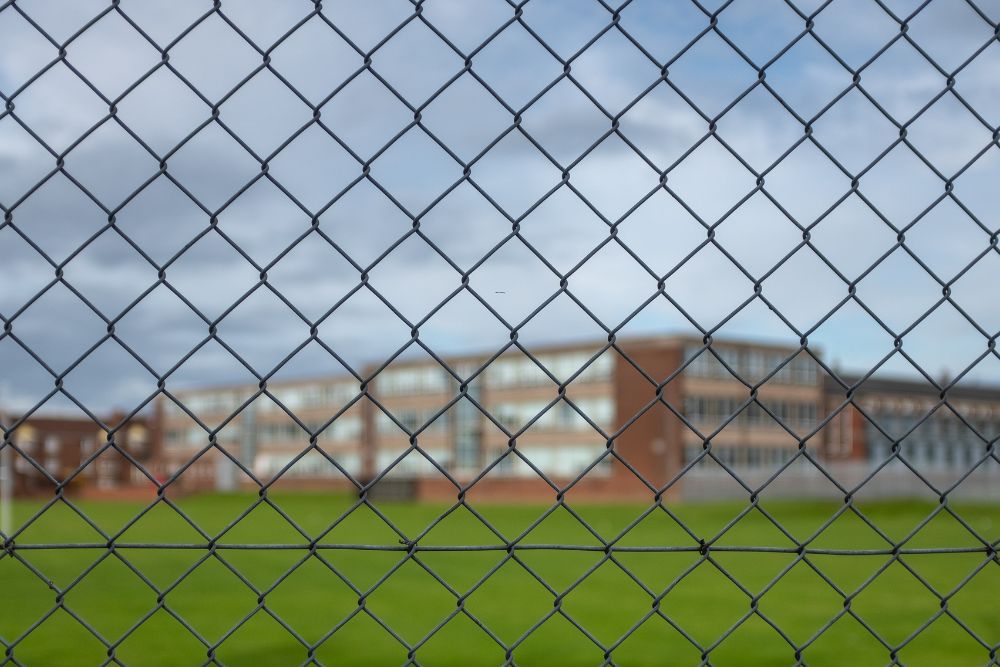 Black chainwire fence enclosing grassy school grounds with a brick school building in the background